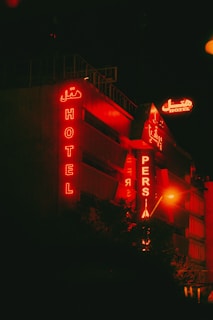 A building with red neon signs displaying the words 'HOTEL' and 'PERSIA' against a dark nighttime backdrop. The signs are both in English and a script which appears to be Persian. The building exterior has a modern design, prominently lit by the red glow from the signage.