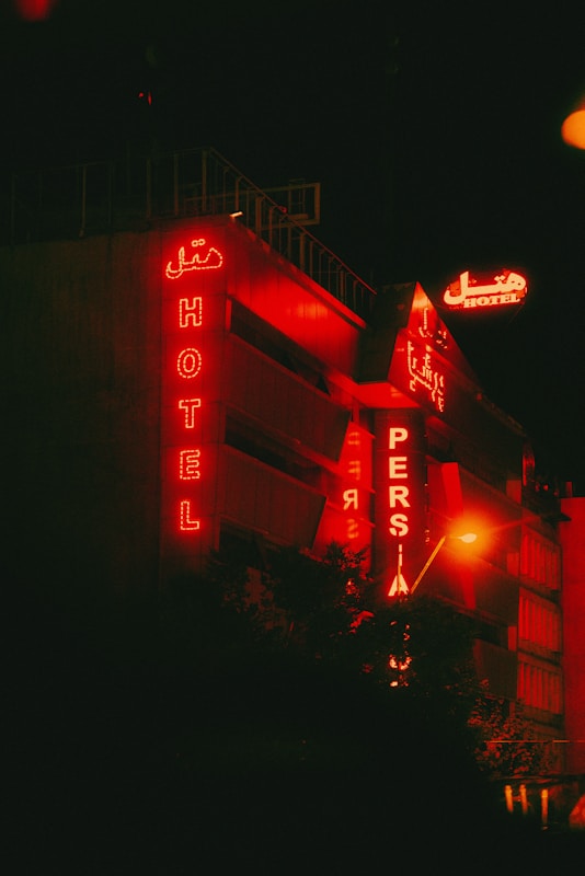 A building with red neon signs displaying the words 'HOTEL' and 'PERSIA' against a dark nighttime backdrop. The signs are both in English and a script which appears to be Persian. The building exterior has a modern design, prominently lit by the red glow from the signage.