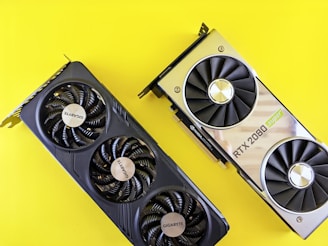 Two graphics cards are placed against a bright yellow background. The card on the left is a Gigabyte model with three fans, featuring a sleek, black design. The card on the right is an RTX 2080 Super, with a metallic finish and dual fans.