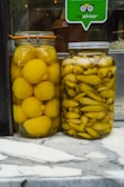 Two jars of preserved vegetables are placed side by side on a marble countertop. The jar on the left contains whole yellow lemons, while the jar on the right contains yellow pickled peppers. Above the jars, there is a TripAdvisor sticker on the glass, indicating a place of business.