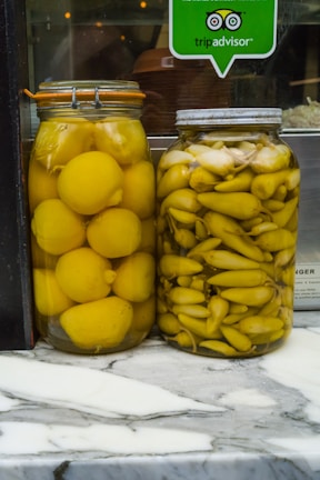 Two jars of preserved vegetables are placed side by side on a marble countertop. The jar on the left contains whole yellow lemons, while the jar on the right contains yellow pickled peppers. Above the jars, there is a TripAdvisor sticker on the glass, indicating a place of business.