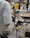 A person is working in an industrial setting, using a power tool that emits sparks. The area is cluttered with various tools and equipment, including a drill and wrenches, on a workbench. Another person is visible in the background, possibly observing or assisting in the operation.