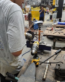A person is working in an industrial setting, using a power tool that emits sparks. The area is cluttered with various tools and equipment, including a drill and wrenches, on a workbench. Another person is visible in the background, possibly observing or assisting in the operation.