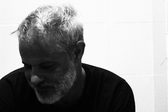 A grayscale portrait of an older man with short, textured hair and a beard, wearing a dark shirt. The man seems to be looking down with a gentle expression, possibly conveying a sense of contemplation. The background is plain and unadorned.