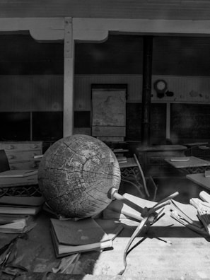 An old classroom setting with a large, weathered globe prominently positioned on a desk amidst scattered papers and books. The background shows vintage chalkboards with faint writing, a map hanging on the wall, and a classic clock. The overall scene evokes a sense of nostalgia and history.