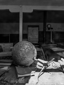 An old classroom setting with a large, weathered globe prominently positioned on a desk amidst scattered papers and books. The background shows vintage chalkboards with faint writing, a map hanging on the wall, and a classic clock. The overall scene evokes a sense of nostalgia and history.
