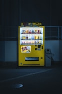 A brightly lit yellow vending machine is set against a dimly lit urban backdrop. It is filled with various beverages, and colorful advertisements are displayed prominently on top. The machine stands next to a waste bin, casting a soft glow on the pavement below.