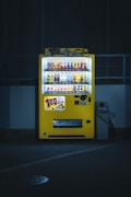A brightly lit yellow vending machine is set against a dimly lit urban backdrop. It is filled with various beverages, and colorful advertisements are displayed prominently on top. The machine stands next to a waste bin, casting a soft glow on the pavement below.