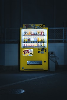 A brightly lit yellow vending machine is set against a dimly lit urban backdrop. It is filled with various beverages, and colorful advertisements are displayed prominently on top. The machine stands next to a waste bin, casting a soft glow on the pavement below.