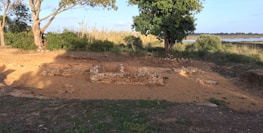 A serene landscape featuring ancient stone foundations partially embedded in the earth. The site is surrounded by trees, shrubs, and a grassy foreground. A light blue sky and a distant water body enhance the natural setting.