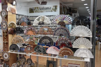 A window display features an array of intricately designed hand fans, each with unique patterns and colors. The fans are arranged in rows on shelves, with the background of a shop interior visible through the glass. Additional decorative objects like clocks and crosses are also displayed on the side.