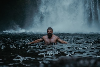 a man in a body of water in front of a waterfall in touch with his male instincts