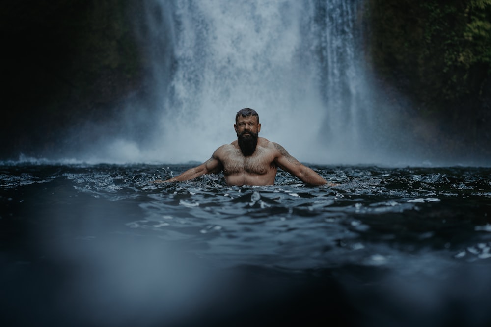 Ein Mann mit Bart sitzt im Wasser vor einem Wasserfall