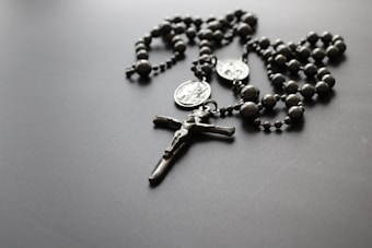 A silver-toned rosary with beaded segments and two medallions lies on a flat, dark surface. The center features a crucifix with a detailed, small depiction of Jesus Christ.