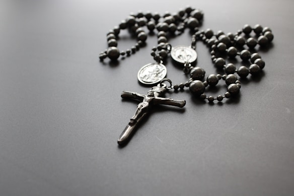A silver-toned rosary with beaded segments and two medallions lies on a flat, dark surface. The center features a crucifix with a detailed, small depiction of Jesus Christ.