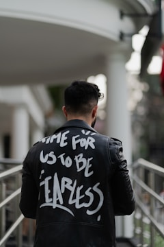 A person wearing a black leather jacket with bold white text that reads 'TIME FOR US TO BE FEARLESS.' stands indoors against a blurred background with columns and metal railings.
