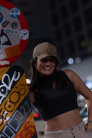 A woman is smiling while standing next to an urban object covered in graffiti and stickers. She is wearing a beige cap and hoop earrings, with a sleeveless black top and light-colored pants. The background is blurred, with some city lights visible, suggesting a nighttime scene.