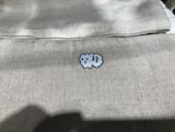 A light-colored fabric features a small, embroidered cartoon-like sheep in the center. The texture of the fabric is visible, giving a sense of softness and simplicity.