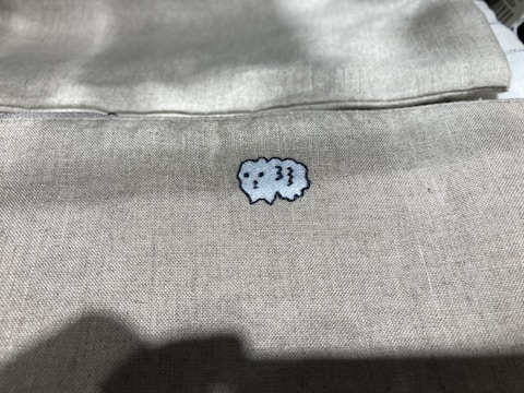 A light-colored fabric features a small, embroidered cartoon-like sheep in the center. The texture of the fabric is visible, giving a sense of softness and simplicity.