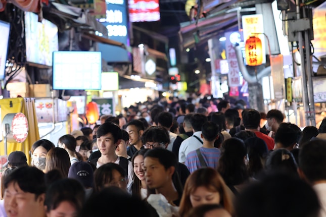 A bustling and crowded street market filled with people walking closely together. Bright and colorful neon signs illuminate the scene, creating a lively atmosphere. Stalls line the sides, displaying various goods and street food.