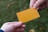 Two hands are exchanging a plain orange rectangular card against a blurred green background. The card is centrally positioned, held by fingers from each hand.