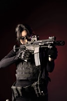 A person wearing tactical gear and dark clothing holds a rifle in a ready position. The individual appears focused, with hair pulled back and wearing protective eyewear. The background is dark, adding to the intensity of the image.