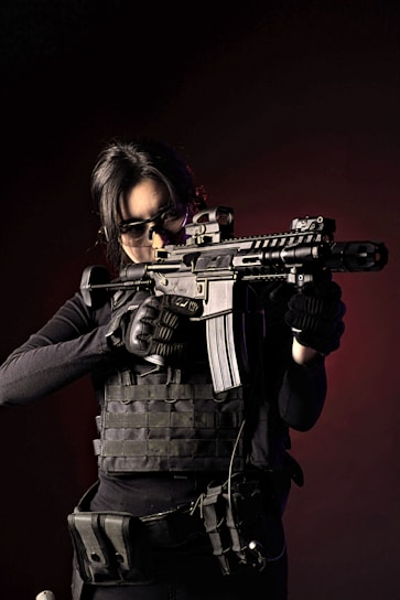 A person wearing tactical gear and dark clothing holds a rifle in a ready position. The individual appears focused, with hair pulled back and wearing protective eyewear. The background is dark, adding to the intensity of the image.