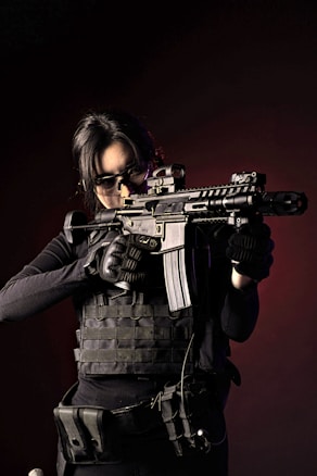 A person wearing tactical gear and dark clothing holds a rifle in a ready position. The individual appears focused, with hair pulled back and wearing protective eyewear. The background is dark, adding to the intensity of the image.