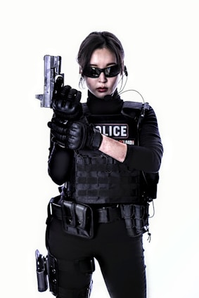 A person dressed in tactical police gear is wearing sunglasses and holding a handgun. The attire includes a black tactical vest, gloves, and various equipment attached to the belt. The expression appears serious and focused.
