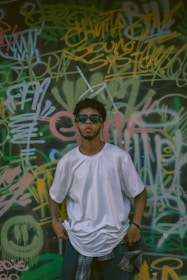 A person wearing a white t-shirt and sunglasses stands in front of a wall covered with colorful graffiti and layered text. The individual has a relaxed pose, with hands in pockets, and a plaid shirt tied around the waist.
