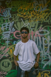 A person wearing a white t-shirt and sunglasses stands in front of a wall covered with colorful graffiti and layered text. The individual has a relaxed pose, with hands in pockets, and a plaid shirt tied around the waist.