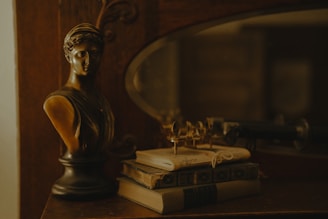 A bronze bust of a classical figure is placed on a wooden surface beside a stack of vintage books. An antique pair of eyeglasses rests on top of the books. The setting appears dimly lit, creating a warm, nostalgic atmosphere with rich, earthy tones.