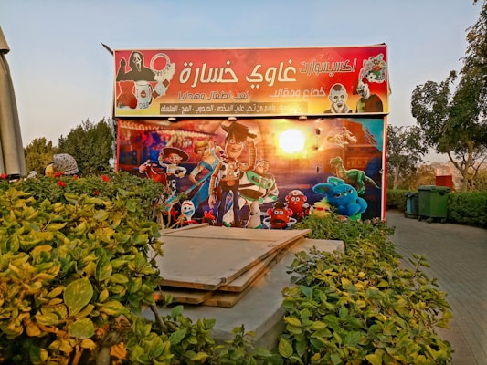 A colorful outdoor advertisement features animated characters, including cowboys, astronauts, and various toys. The sign is decorated with vibrant graphics, and there are lush green plants and a walkway near it.