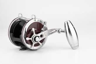 A metallic fishing reel with a polished, shiny surface. It features a crank handle and precision-engineered components, combining both functionality and aesthetics. The reel has an elegant and modern design.