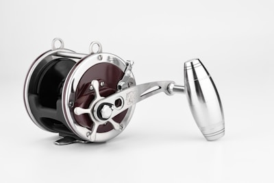 A metallic fishing reel with a polished, shiny surface. It features a crank handle and precision-engineered components, combining both functionality and aesthetics. The reel has an elegant and modern design.