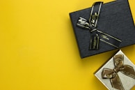 Two gift boxes with ribbons are placed on a bright yellow background. The upper box is black with a black ribbon, while the lower box is silver with a brown ribbon.