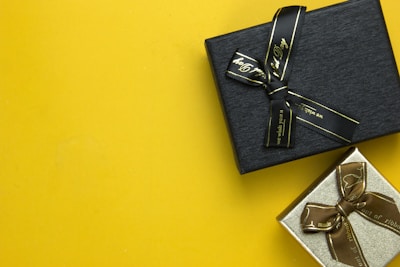 Two gift boxes with ribbons are placed on a bright yellow background. The upper box is black with a black ribbon, while the lower box is silver with a brown ribbon.
