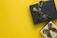 Two gift boxes with ribbons are placed on a bright yellow background. The upper box is black with a black ribbon, while the lower box is silver with a brown ribbon.
