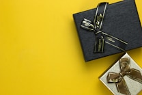 Two gift boxes with ribbons are placed on a bright yellow background. The upper box is black with a black ribbon, while the lower box is silver with a brown ribbon.