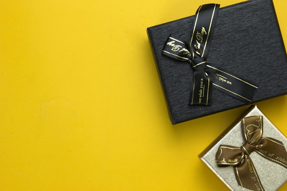 Two gift boxes with ribbons are placed on a bright yellow background. The upper box is black with a black ribbon, while the lower box is silver with a brown ribbon.