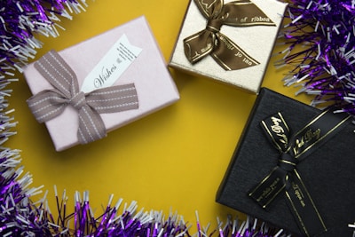 Three gift boxes are arranged on a yellow surface. The boxes are adorned with bows, each in a different color: pink, beige, and black. Tinsel surrounds the setup, featuring purple and white strands, adding an element of festivity.
