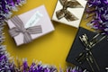 Three gift boxes are arranged on a yellow surface. The boxes are adorned with bows, each in a different color: pink, beige, and black. Tinsel surrounds the setup, featuring purple and white strands, adding an element of festivity.