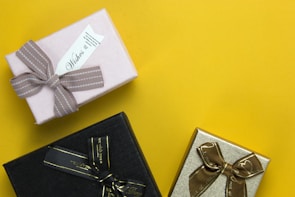 Three neatly wrapped gift boxes are arranged on a vibrant yellow background. One box is wrapped in light pink paper with a textured taupe ribbon and a tag reading 'Wishes.' The second box is wrapped in black paper with a glossy black ribbon featuring gold accents. The third box is wrapped in shimmering gold paper with a brown ribbon decorated with gold details.