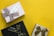 Three neatly wrapped gift boxes are arranged on a vibrant yellow background. One box is wrapped in light pink paper with a textured taupe ribbon and a tag reading 'Wishes.' The second box is wrapped in black paper with a glossy black ribbon featuring gold accents. The third box is wrapped in shimmering gold paper with a brown ribbon decorated with gold details.