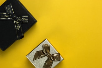 a black box with a gold ribbon and a silver box with a brown bow