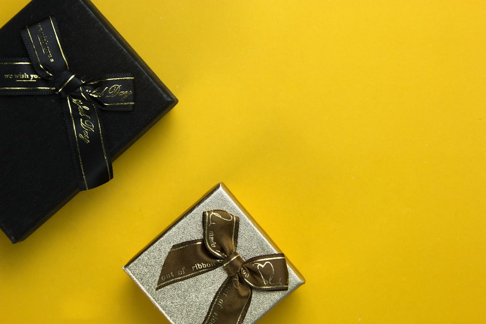 a black box with a gold ribbon and a silver box with a brown bow
