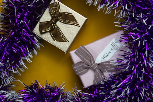 Two wrapped gift boxes are placed on a vibrant yellow surface. One box is gold with a brown ribbon, while the other is pink with a taupe ribbon. The presents are surrounded by a garland of purple and white tinsel.