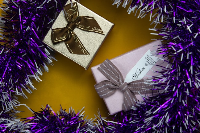 Two wrapped gift boxes are placed on a vibrant yellow surface. One box is gold with a brown ribbon, while the other is pink with a taupe ribbon. The presents are surrounded by a garland of purple and white tinsel.
