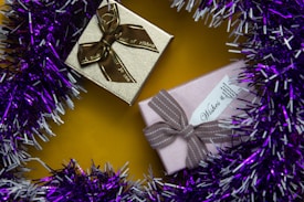 Two wrapped gift boxes are placed on a vibrant yellow surface. One box is gold with a brown ribbon, while the other is pink with a taupe ribbon. The presents are surrounded by a garland of purple and white tinsel.
