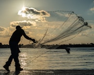 A silhouetted figure is casting a large fishing net over a calm body of water as the sun sets in the background. The sky is adorned with scattered clouds, and a hint of orange hues reflects on the water's surface. In the distance, the outline of trees and buildings can be seen along the horizon.